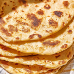 Freshly cooked yogurt flatbreads stacked on a wooden board with olive oil for dipping.