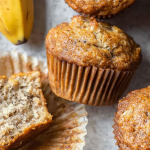 Freshly baked banana muffins on a wooden table, topped with powdered sugar and sliced bananas.