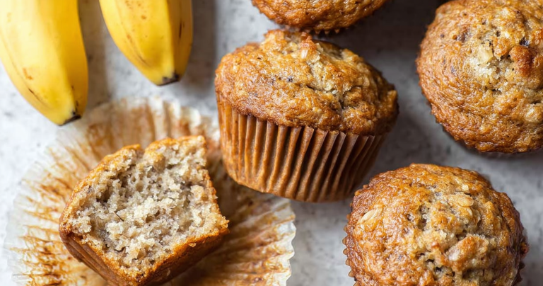 Freshly baked banana muffins on a wooden table, topped with powdered sugar and sliced bananas.