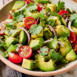 A colorful avocado salad bowl with cherry tomatoes, cucumber, and cilantro on a wooden table.