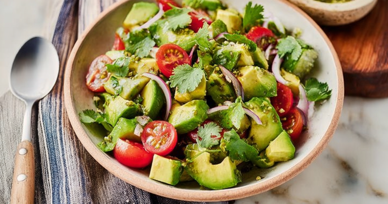 A colorful avocado salad bowl with cherry tomatoes, cucumber, and cilantro on a wooden table.