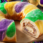 Festively adorned king cake on a table with Mardi Gras decorations, featuring vibrant purple, green, and gold sugars.