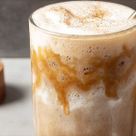 Banana bread latte served in a glass with ice, topped with whipped cream and cinnamon, surrounded by fresh bananas and a slice of banana bread.