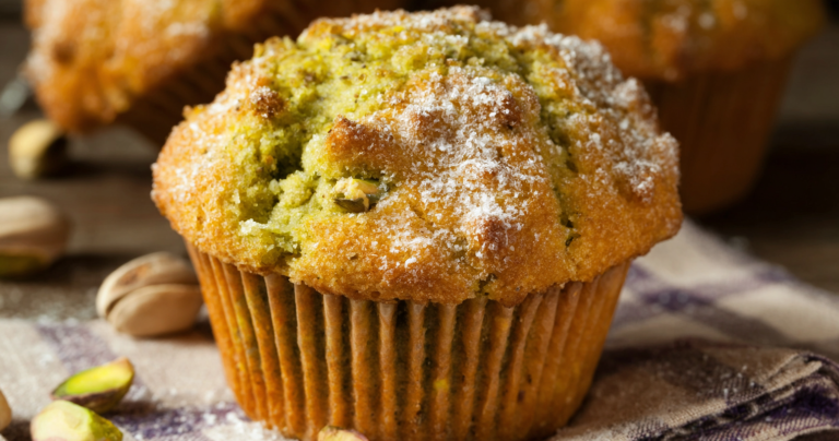 A close-up view of soft and fluffy pistachio muffins on a wooden table with shelled pistachios and a green napkin.