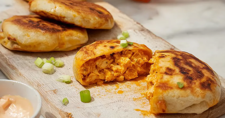 Golden-brown buffalo chicken hot pockets with chives on a wooden table, served with a bowl of dipping sauce.