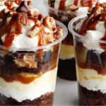 Loaded Brownie Cheesecake Cups with chocolate ganache and caramel drizzle on a wooden table.