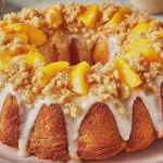 Slice of peach cobbler pound cake with peach preserves and crumble on a white plate, garnished with fresh peaches and mint leaves.