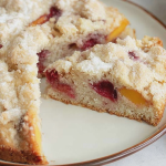 Slice of raspberry peach cake on a decorative plate, topped with fresh raspberries and peach slices, on a wooden table.